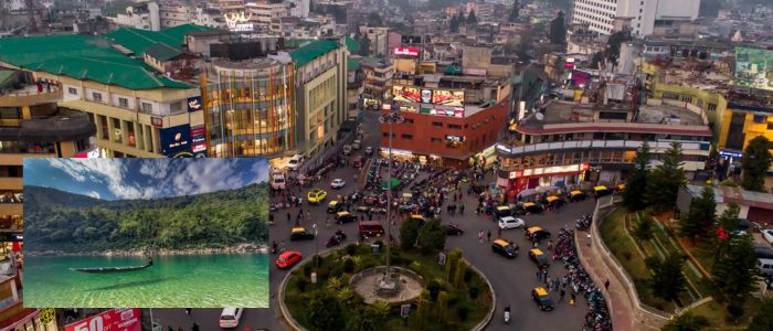 An aerial view of a bustling city center with a roundabout, buildings, and many vehicles. An inset shows a serene green lake with a boat amidst lush mountains.