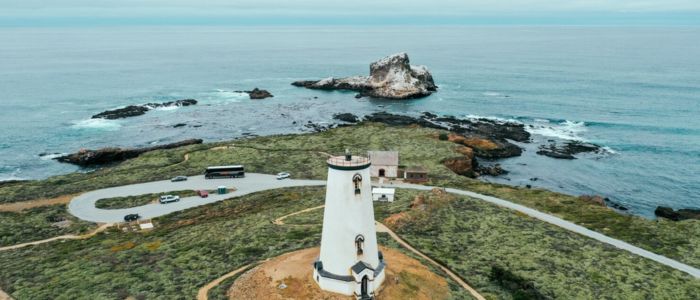 Aerial view of a coastal lighthouse surrounded by waves and rocky shoreline under a clear blue sky.