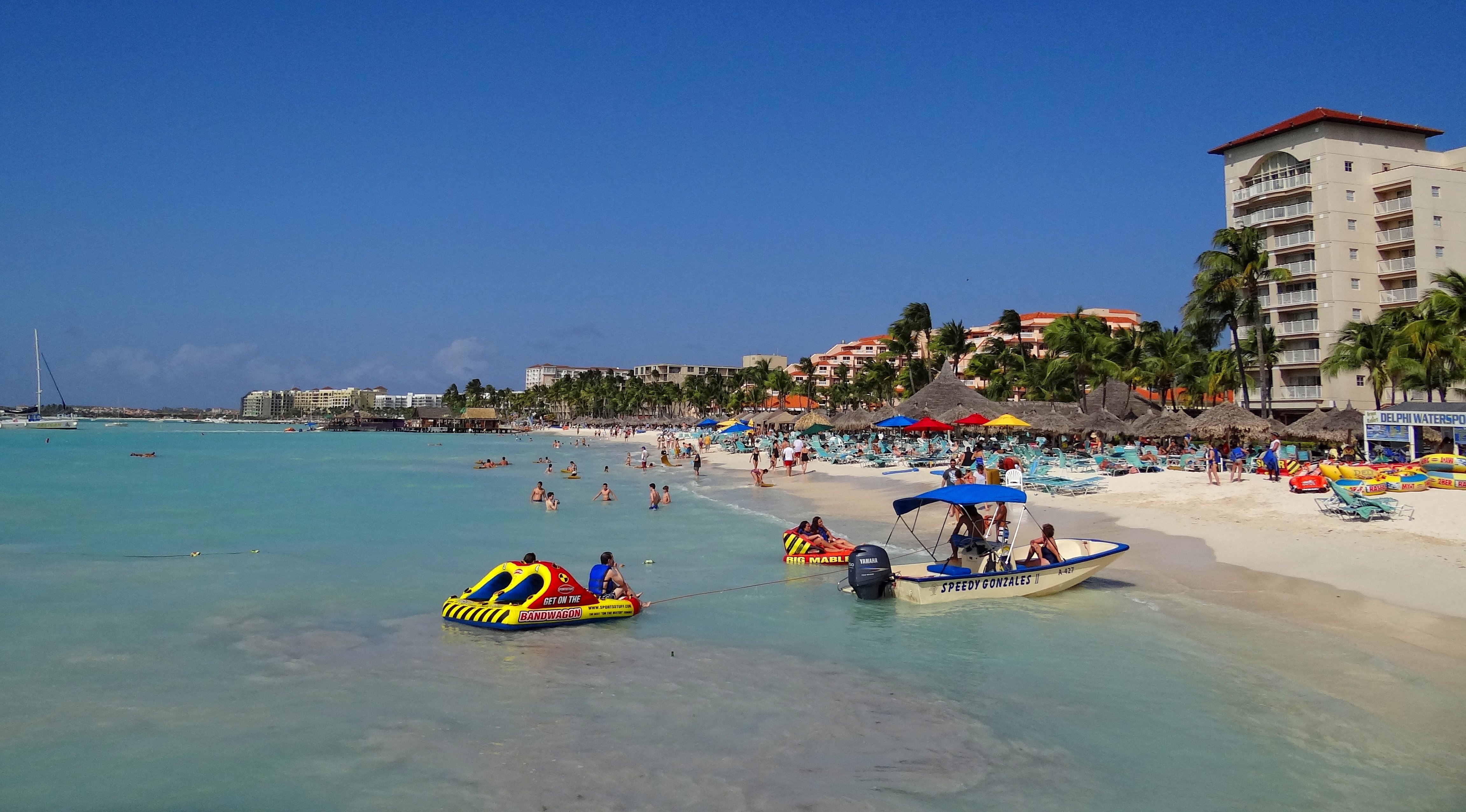 A sunny beach scene with people swimming and boats floating in the clear blue water.