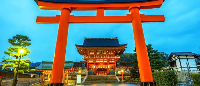 A large red torii gate frames a traditional Japanese temple surrounded by trees at dusk, creating a serene and majestic atmosphere.