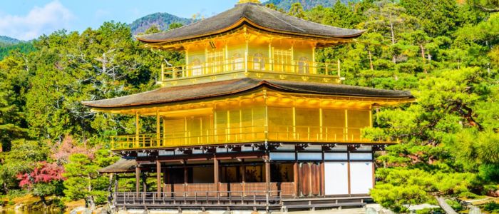 A golden Japanese temple with tiered roofs is set amidst lush greenery and trees, with mountains in the background. The scene conveys tranquility and beauty.