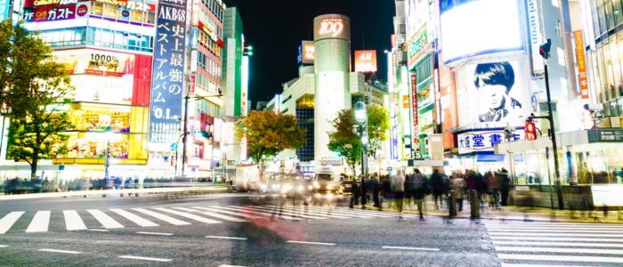 Brightly lit city intersection at night, with blurred pedestrians crossing. Neon signs and video screens on surrounding buildings create a vibrant, bustling atmosphere.