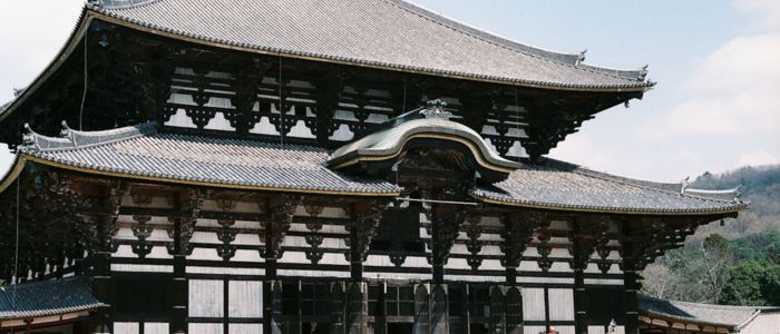 Large traditional Japanese temple with intricate wooden architecture and a tiled roof, set against a backdrop of green trees and a cloudy sky.
