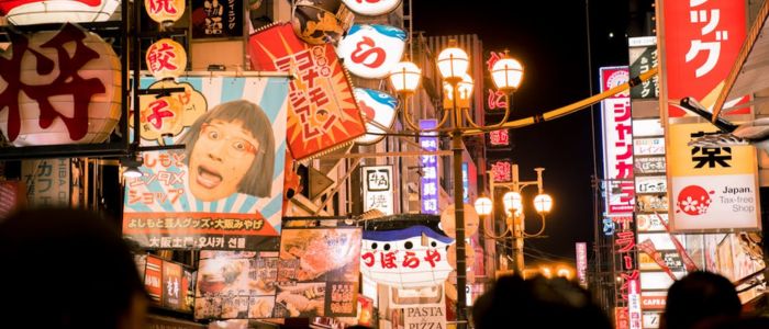 Bustling Japanese street at night, filled with colorful neon signs and lanterns. The scene is vibrant and lively, suggesting a busy urban atmosphere.