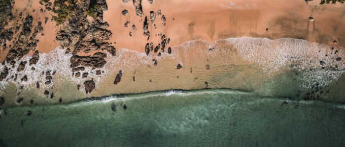 Aerial view of a serene ocean meeting a sandy beach, showcasing waves gently lapping at the shore under a clear sky.