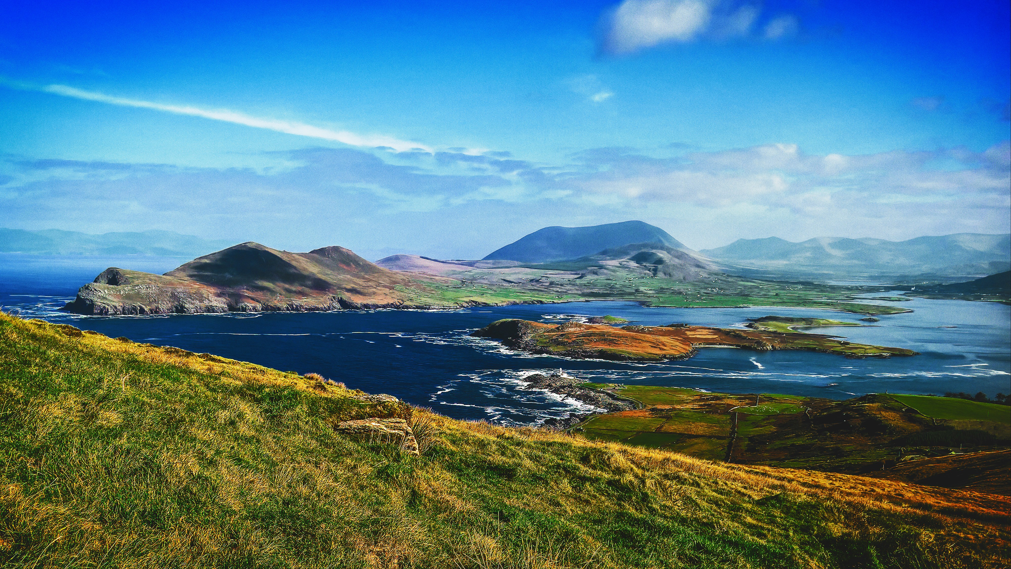 Panoramic view of rugged coastal cliffs and rolling green hills overlooking the Atlantic Ocean under a bright blue sky in Ireland.