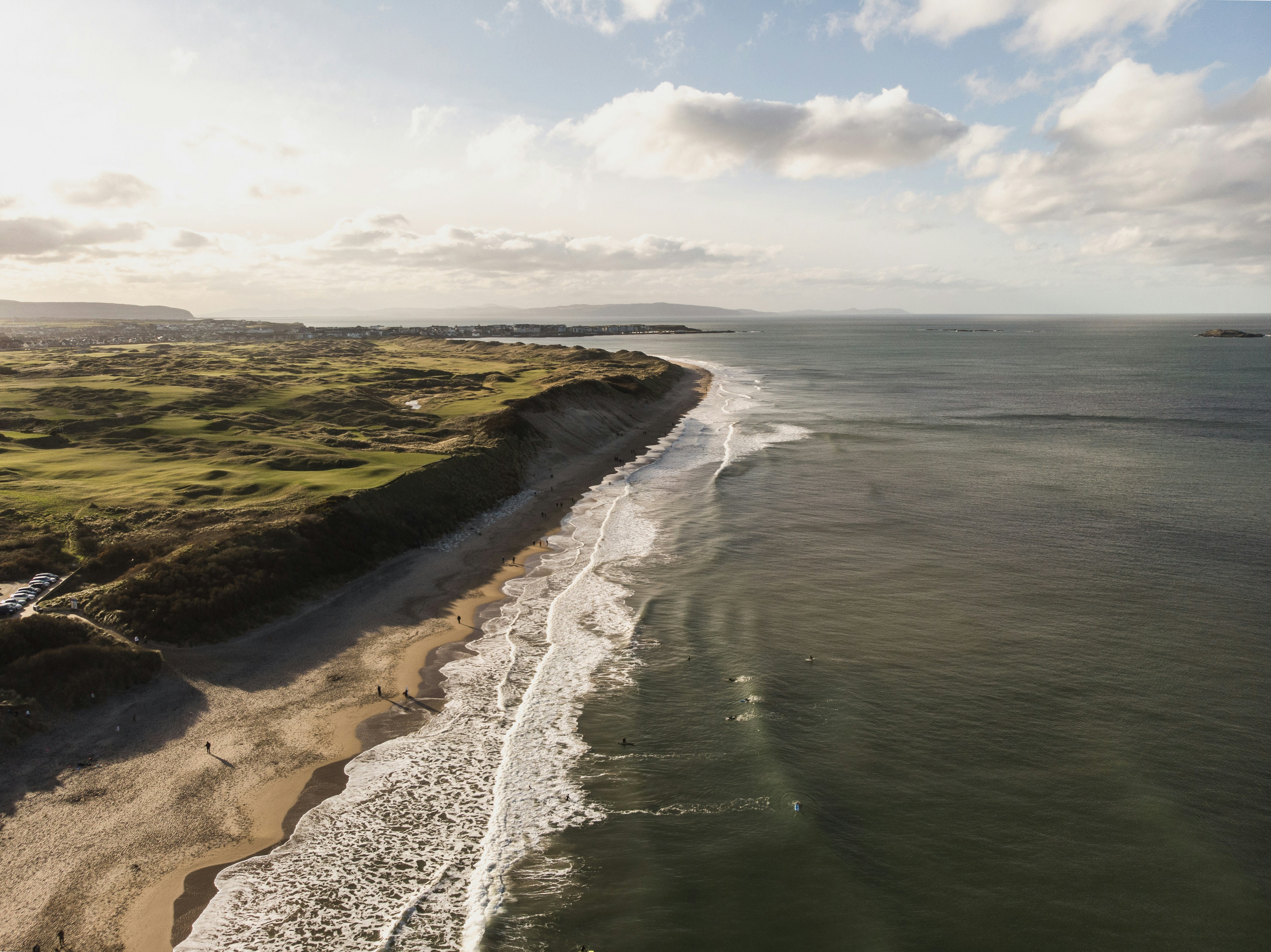 Aerial view of a scenic coastline with sandy beach, rolling green cliffs, and ocean waves under a cloudy sky.